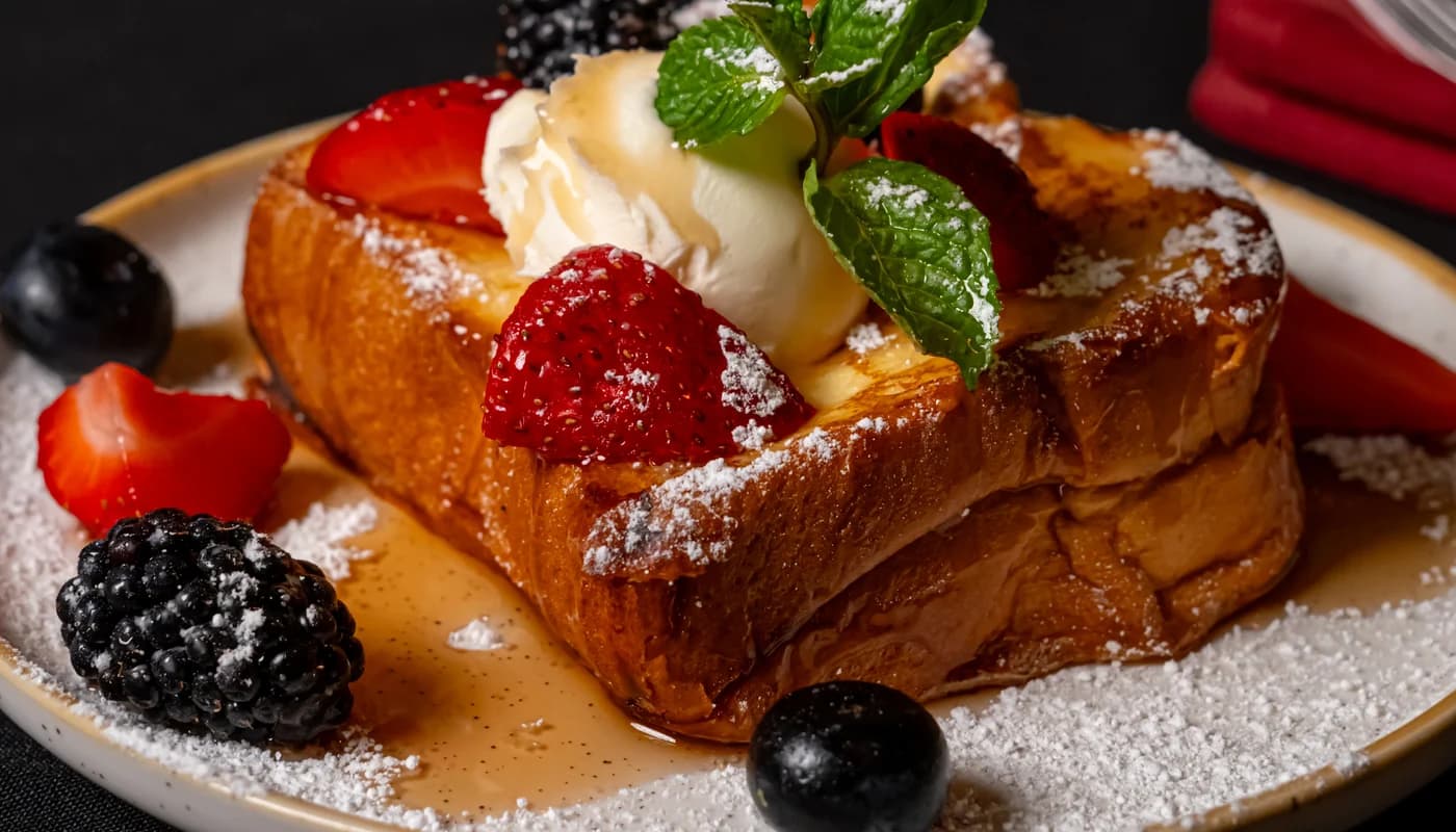 Thick-cut French toast under whipped cream, strawberries, blackberries and mint, dusted with icing sugar on a pale plate.