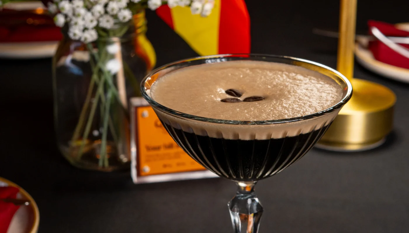 An espresso martini in a coupe with three coffee beans on the foam, white gypsophila, a red rose and a small Spanish flag on the table behind it.