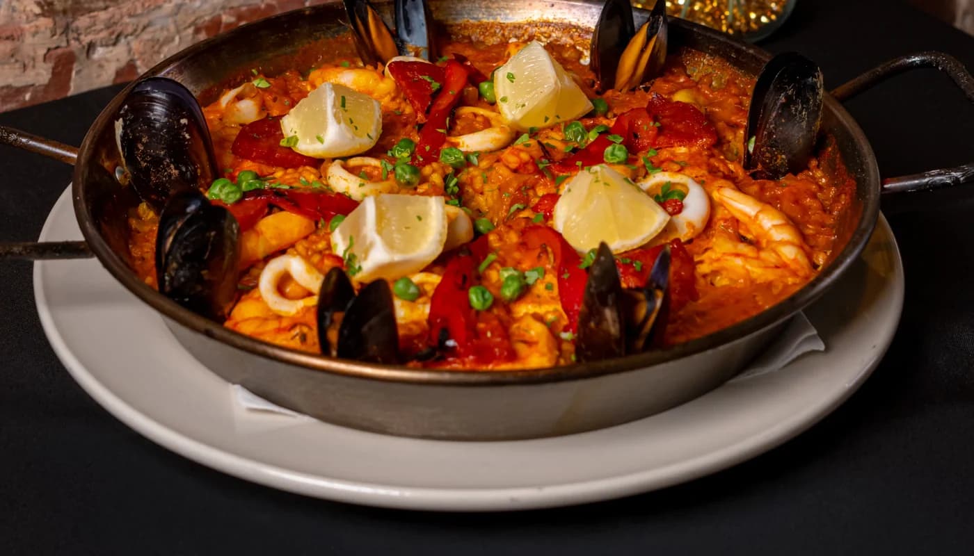 A paella pan of saffron rice with mussels, prawns and lemon wedges, on a table against exposed brick with red roses behind it.