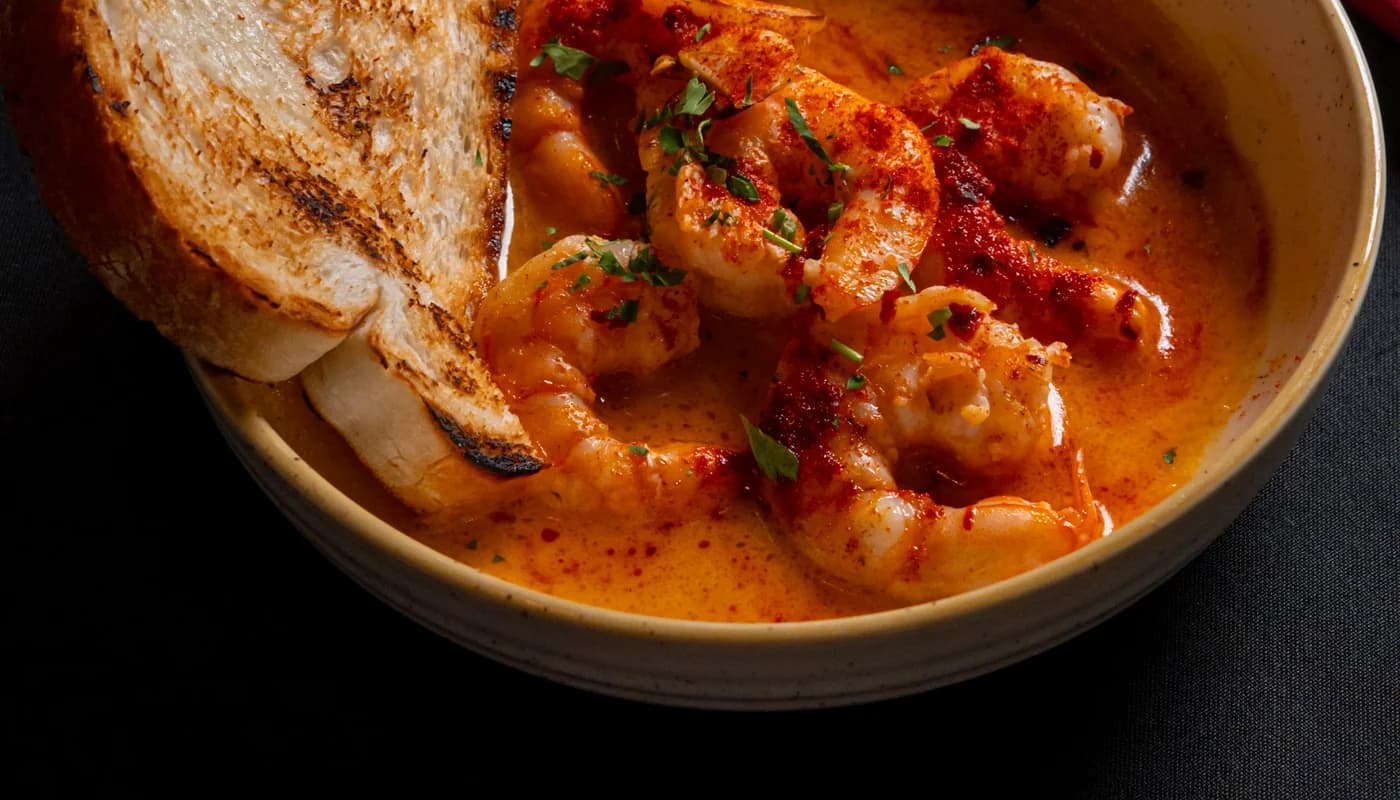 A bowl of gambas al ajillo: prawns in paprika-red garlic sauce with two pieces of grilled bread, and a cocktail on the table behind.
