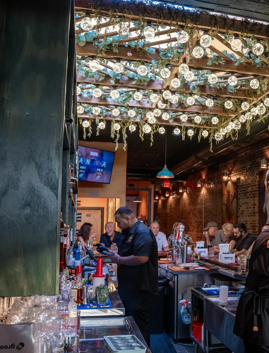The bar at service under filament bulbs strung through eucalyptus: a bartender working the rail, guests along the counter, brick wall behind.