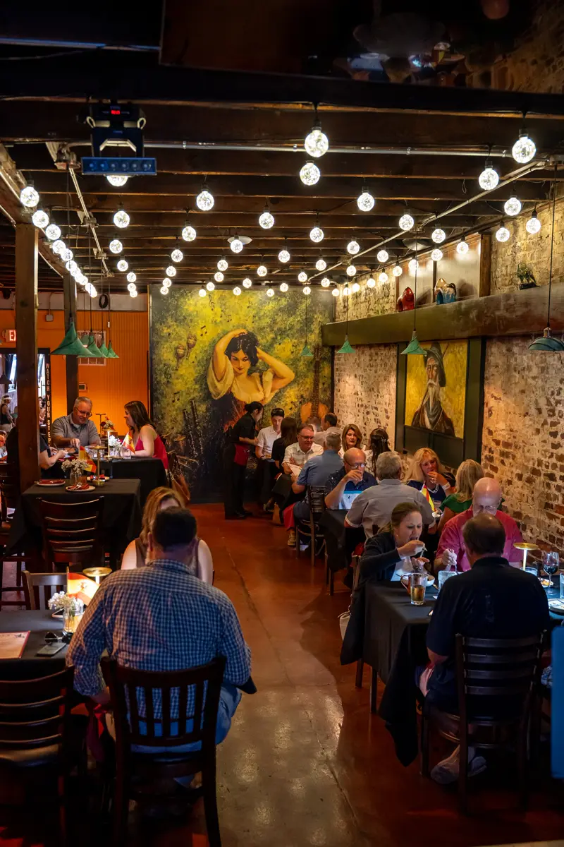 The long dining room at service, tables full along both walls under a canopy of filament bulbs, with painted murals covering the far end.