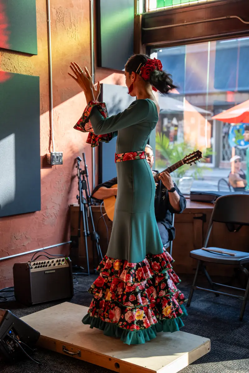 A flamenco dancer in a green ruffled dress stands beside a seated guitarist on the small stage by the window during a live set.