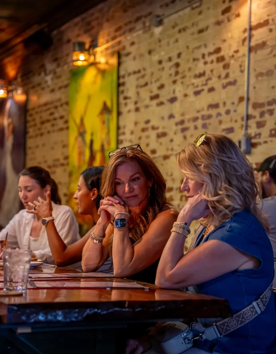 Four guests leaning together over drinks at the timber bar, against the brick wall.