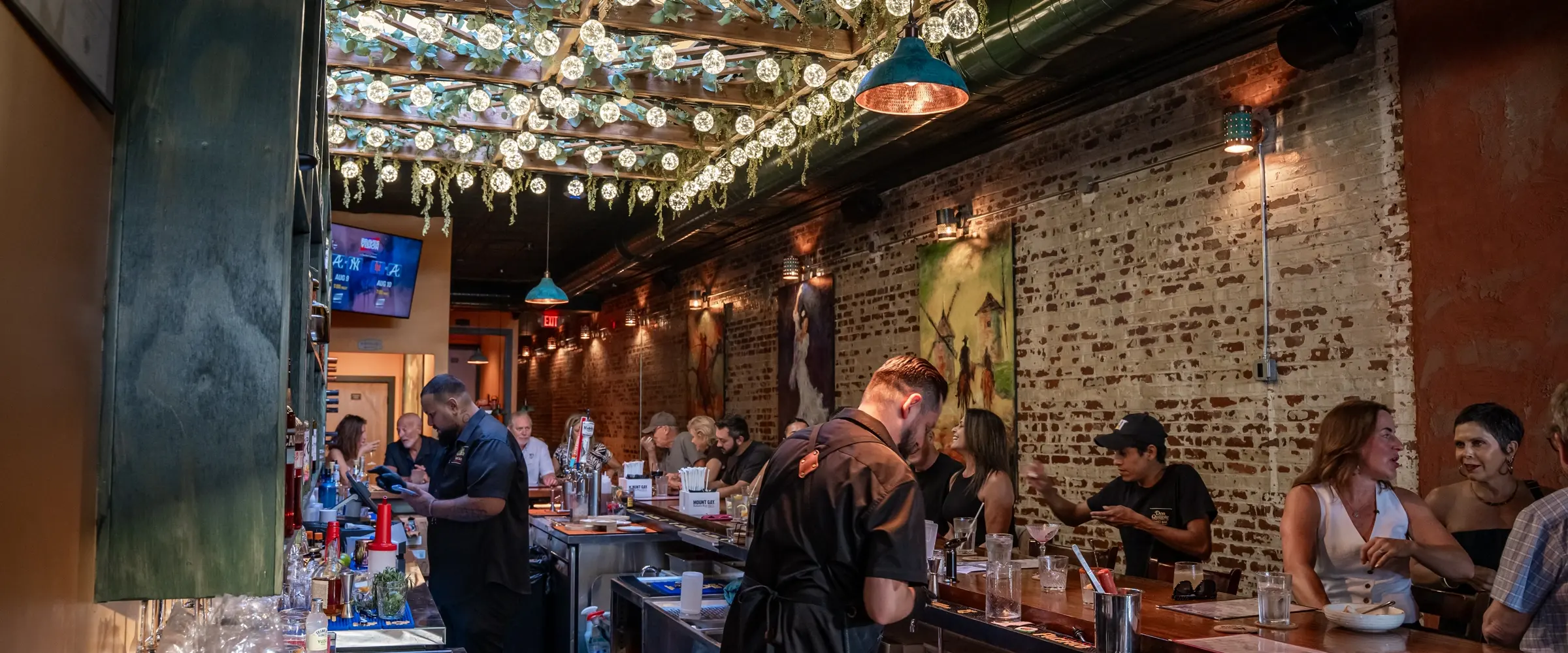 Two cooks working the pass behind the bar under a canopy of filament bulbs, guests seated along the rail.