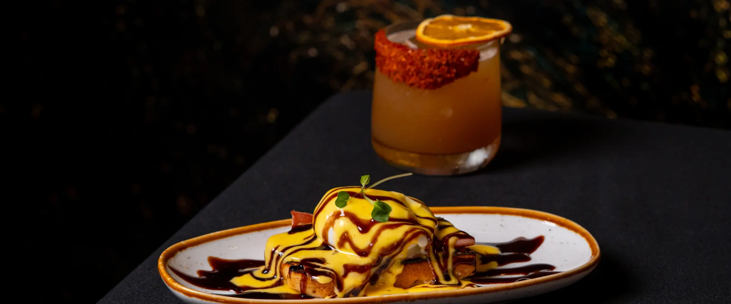 A plate of eggs benedict under saffron sauce and a dark drizzle, with a citrus-garnished cocktail behind it on the black table.