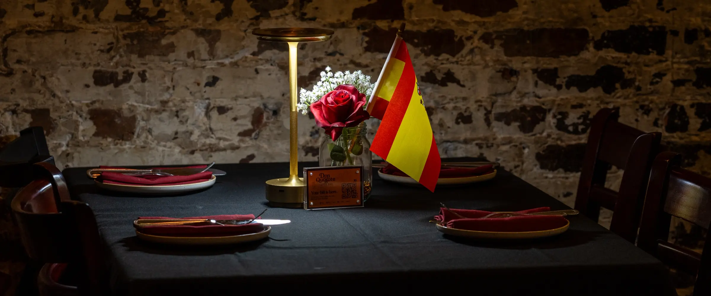 A table laid and waiting against the brick wall: black cloth, red plates and napkins, and a rose and a small Spanish flag standing in a brass holder.