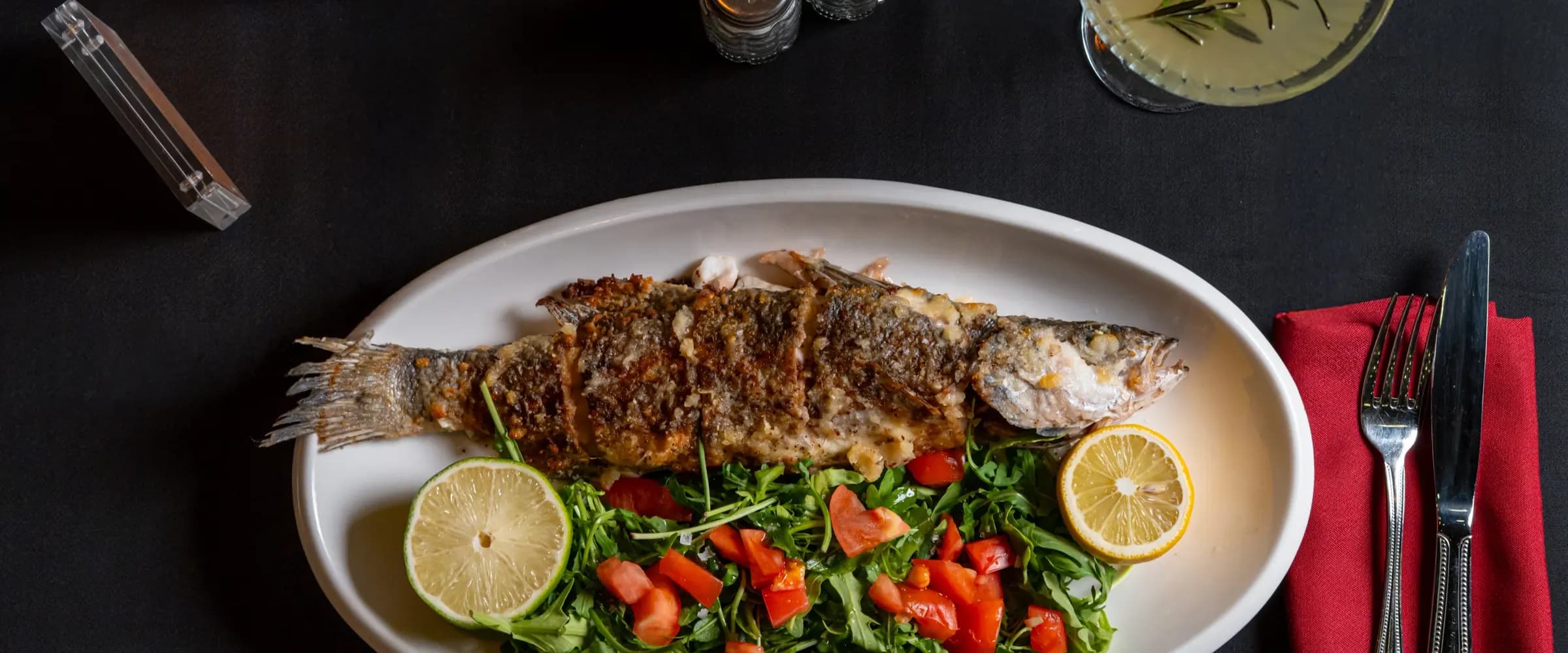 A whole grilled fish on an oval platter over rocket, with diced tomato, lime and lemon, and a herb-garnished cocktail on the table beside it.