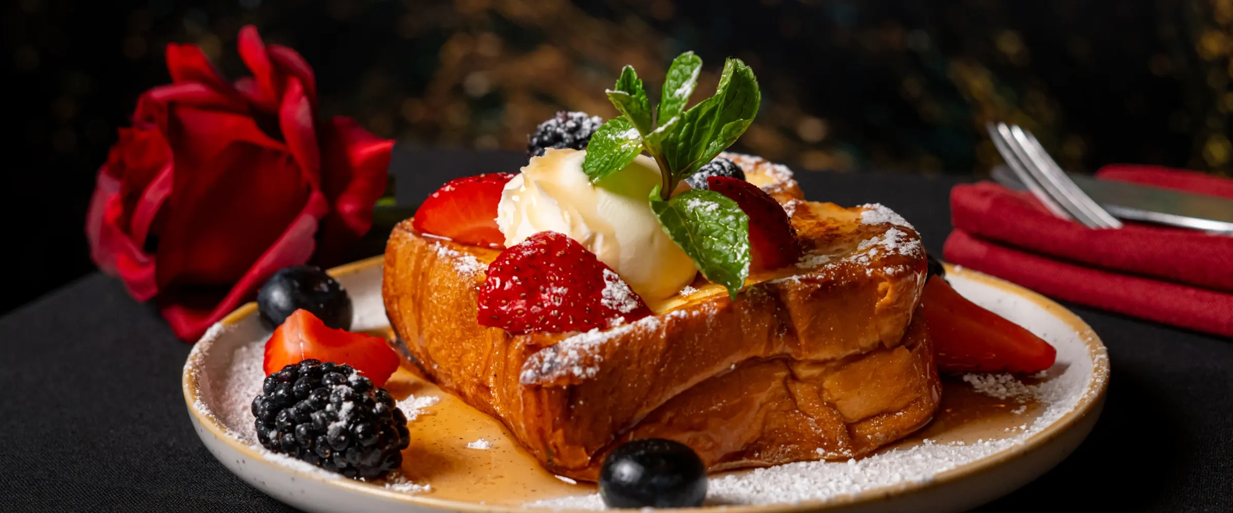 A thick slice of brioche french toast dusted with sugar, topped with cream, mint and berries, with a red rose on the table beside it.
