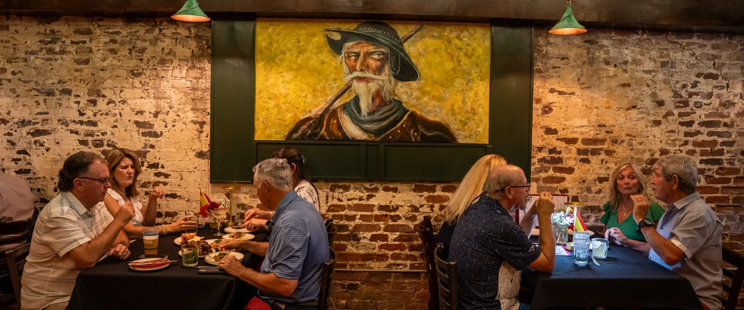 The dining room full at dinner: tables of guests under the painting of Don Quixote, filament bulbs strung along the brick wall.