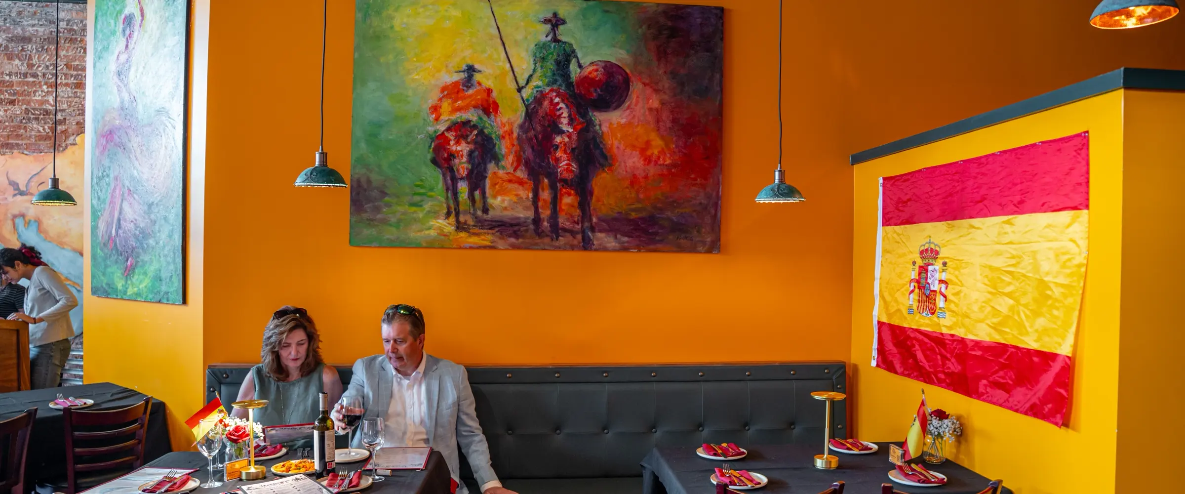The ochre dining room: a large painting of Don Quixote and Sancho on horseback over banquette tables laid with red napkins and flags.